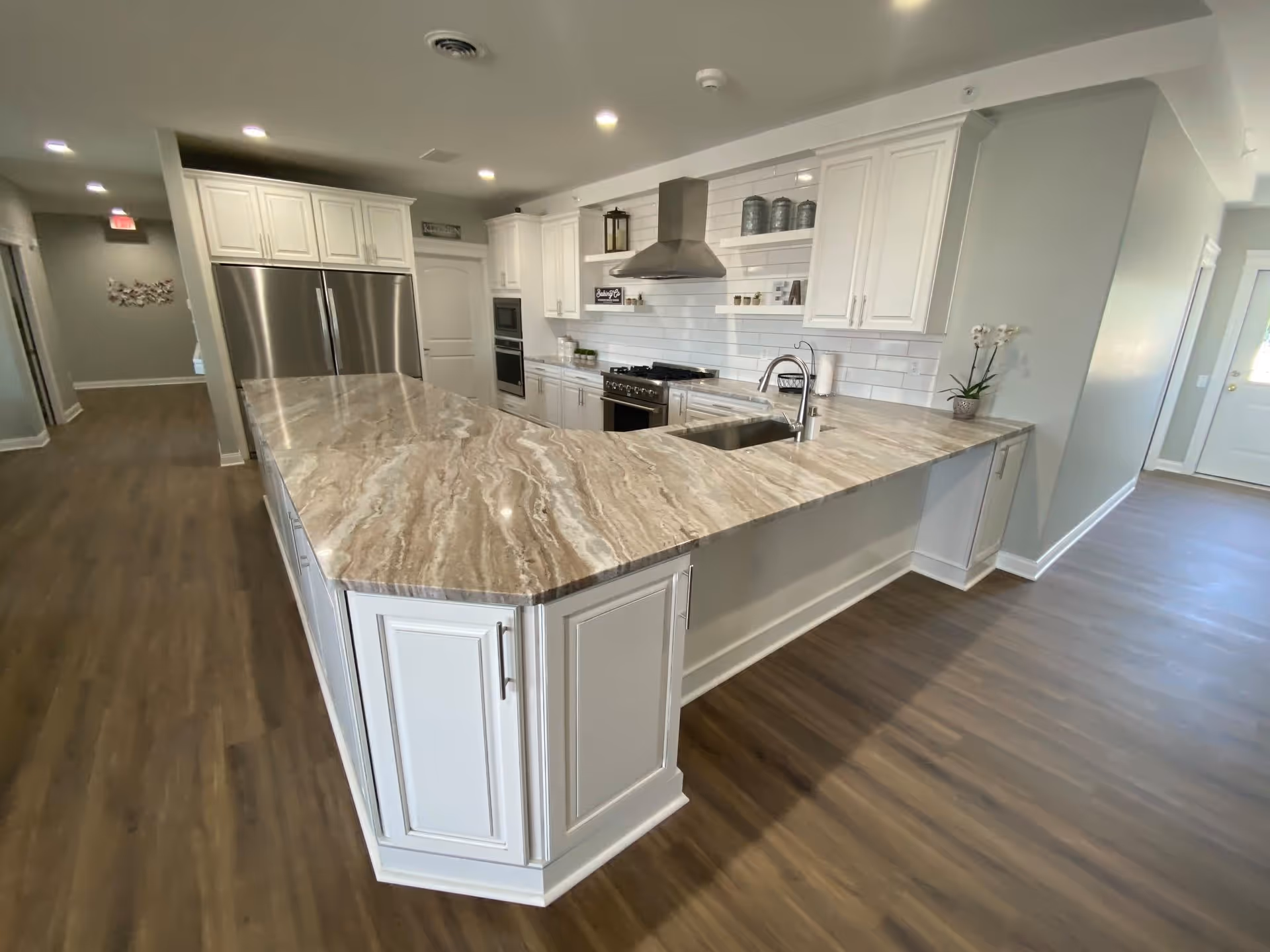 A modern kitchen with a large marble countertop island, white cabinets, stainless steel refrigerator, oven, and range hood. The kitchen has wooden flooring and white subway tile backsplash with open shelves displaying decorative items.