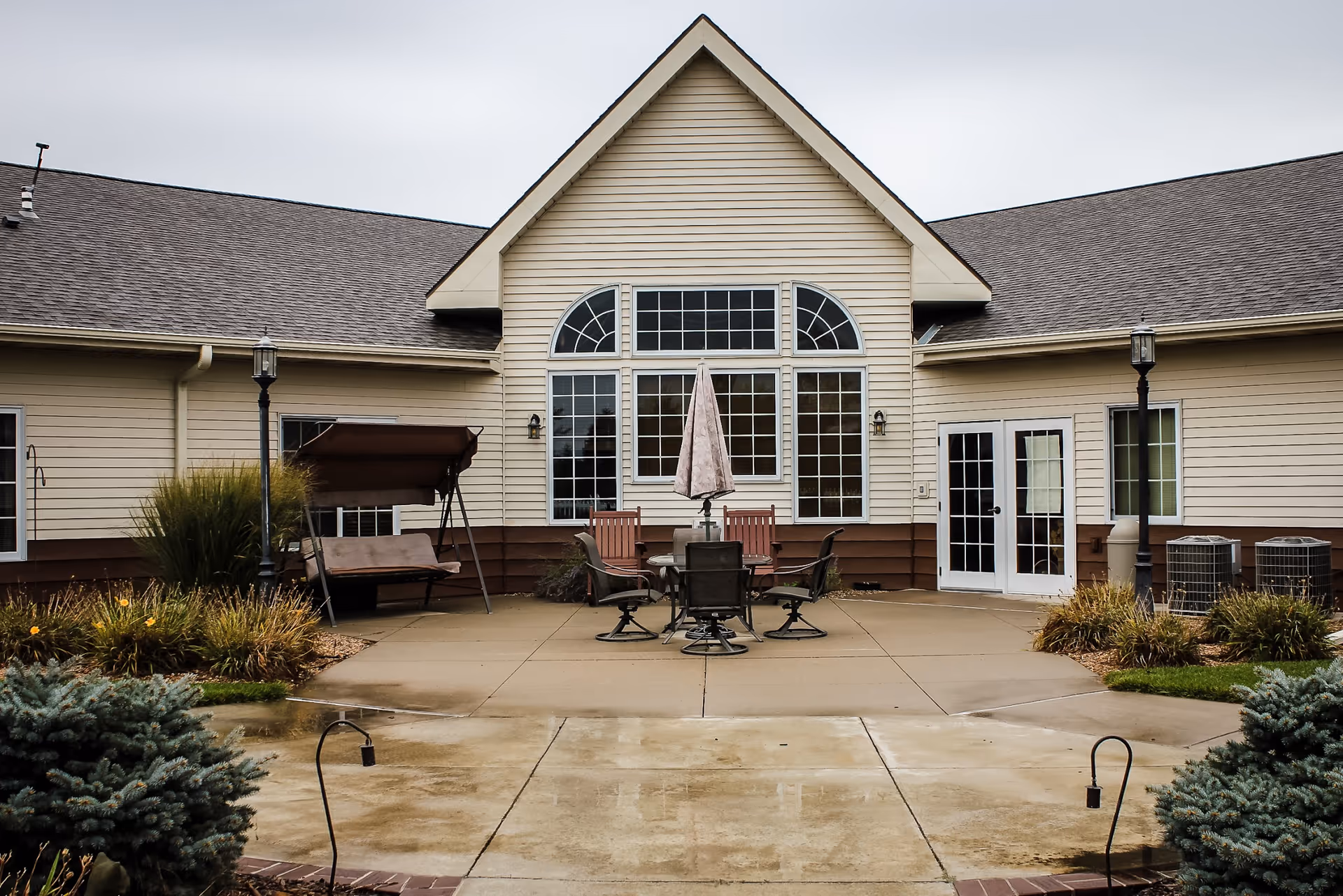 Outdoor patio courtyard with a table, chairs, umbrella and swing in front of a beige senior living building facade.
