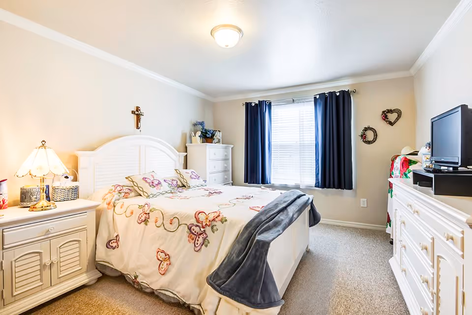 A bright and cozy bedroom with a white wooden bed frame and floral bedding featuring butterfly designs. There is a nightstand with a lamp and decorative items on the left side of the bed. A white dresser with a TV on top is on the right side of the room. The window has dark blue curtains, and the walls are decorated with two heart-shaped wreaths. The room has beige carpet and neutral-colored walls.