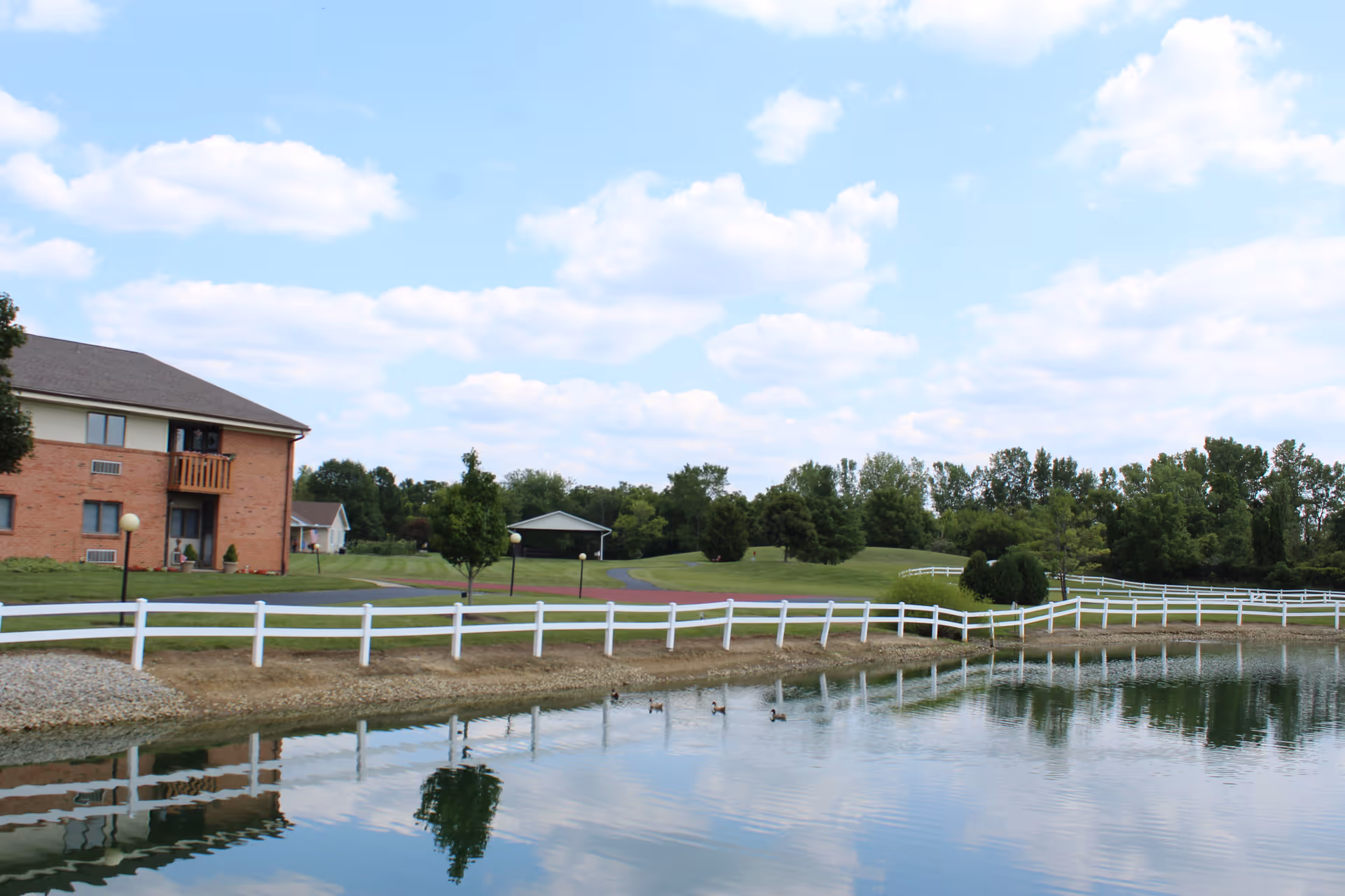 A peaceful outdoor scene at Brookhaven Retirement Community featuring a pond with ducks swimming, a white fence running along the water's edge, a grassy area with trees, and a brick building with balconies on the left side under a partly cloudy sky.