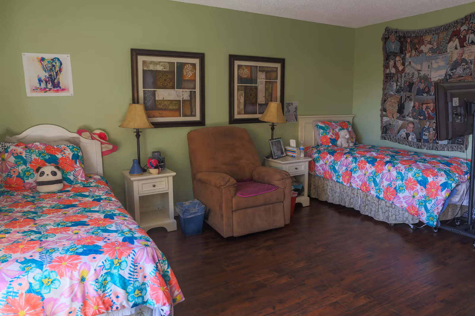 A cozy bedroom with two twin beds featuring colorful floral bedspreads. Between the beds is a brown armchair and two white nightstands with lamps. The walls are painted light green and decorated with framed artwork and a large tapestry. The floor is wooden.