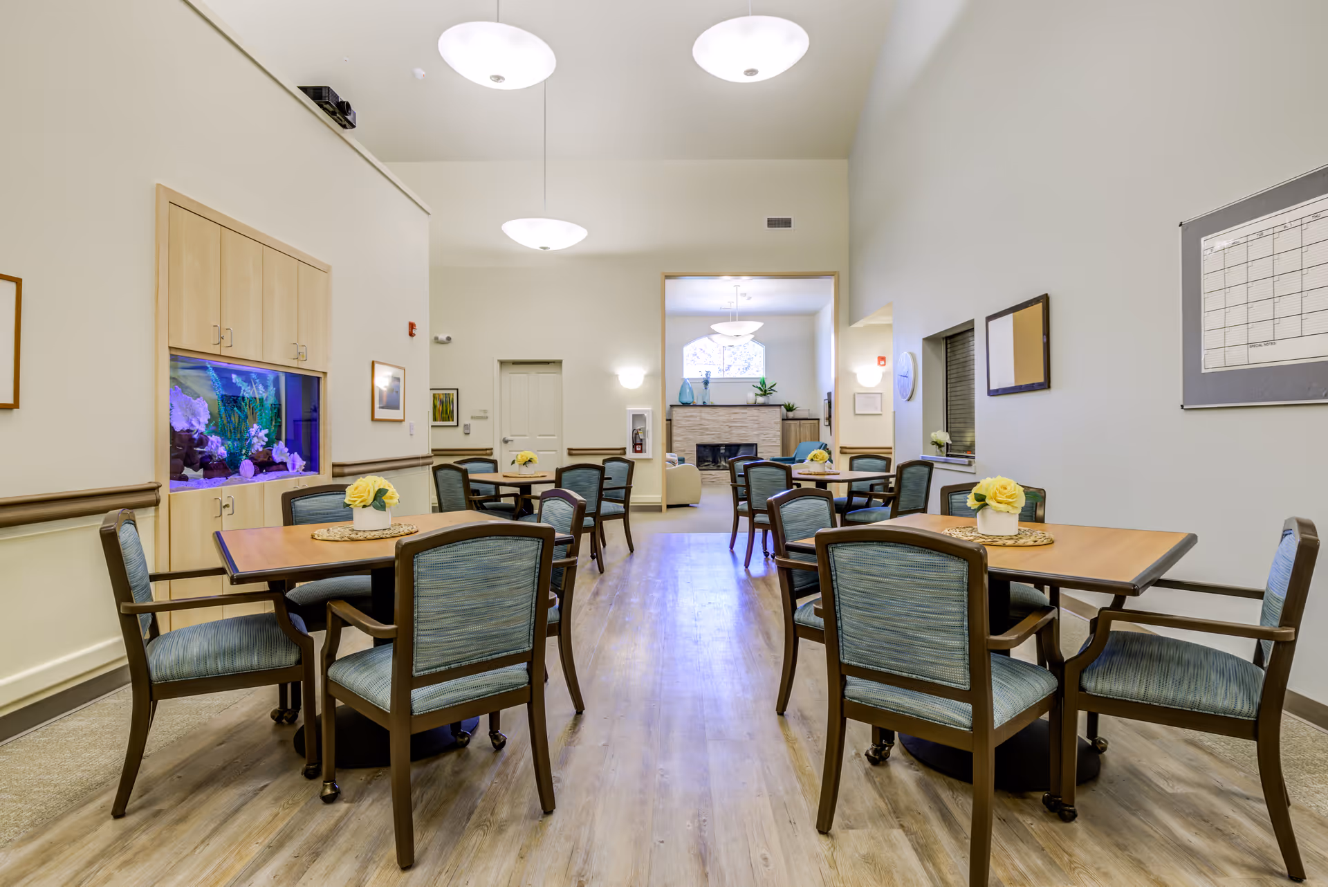 A bright and clean common dining area in a senior living facility with several wooden tables and blue cushioned chairs. Each table has a small flower arrangement. There is a built-in aquarium on the left wall and a cozy lounge area with a fireplace visible through an open doorway at the back. The room has light-colored walls and wood flooring with modern ceiling lights.