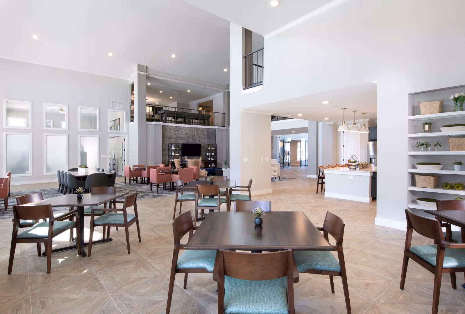 A spacious, modern common area in a senior living facility featuring multiple wooden tables with green cushioned chairs. The room has high ceilings, large windows, and a mix of seating including pink armchairs near a TV mounted on a stone accent wall. There is an open kitchen area with pendant lights and shelves with decorative plants and baskets.