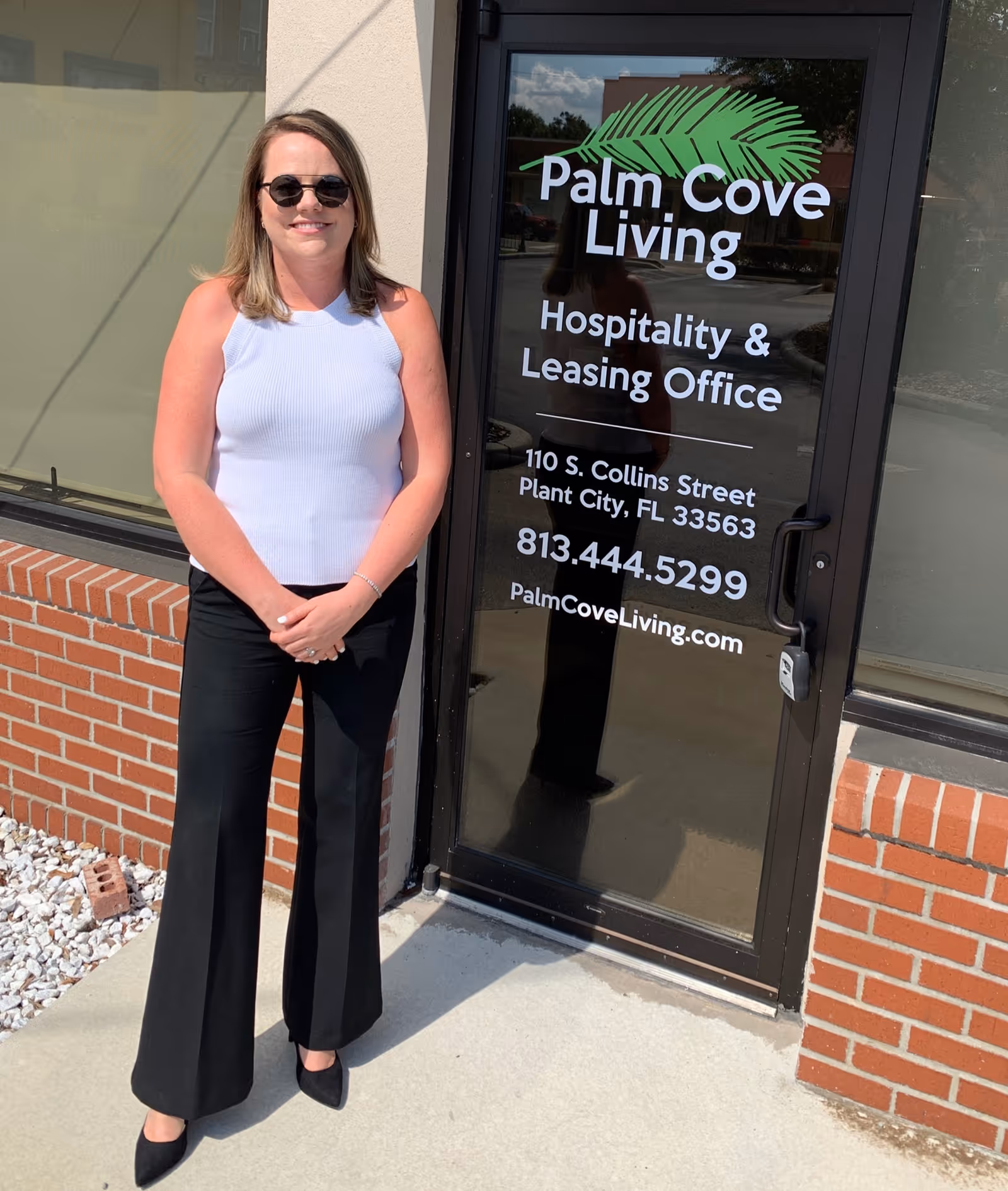 A woman in sunglasses stands outside the Palm Cove Living hospitality and leasing office next to a glass door displaying the facility name, address, and phone number.