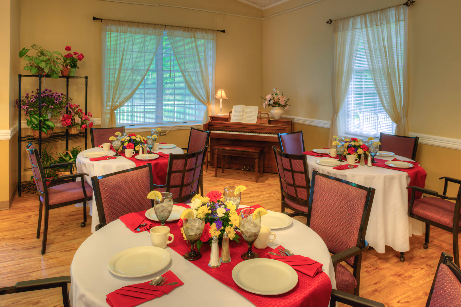 A cozy dining room with round tables covered in white tablecloths and red runners, set with plates, cups, utensils, and glasses with lemon slices. Each table has a floral centerpiece. The room has wooden floors, beige walls, two windows with sheer curtains, a piano with a lamp and flower vase on top, and a black metal plant stand with various potted plants.