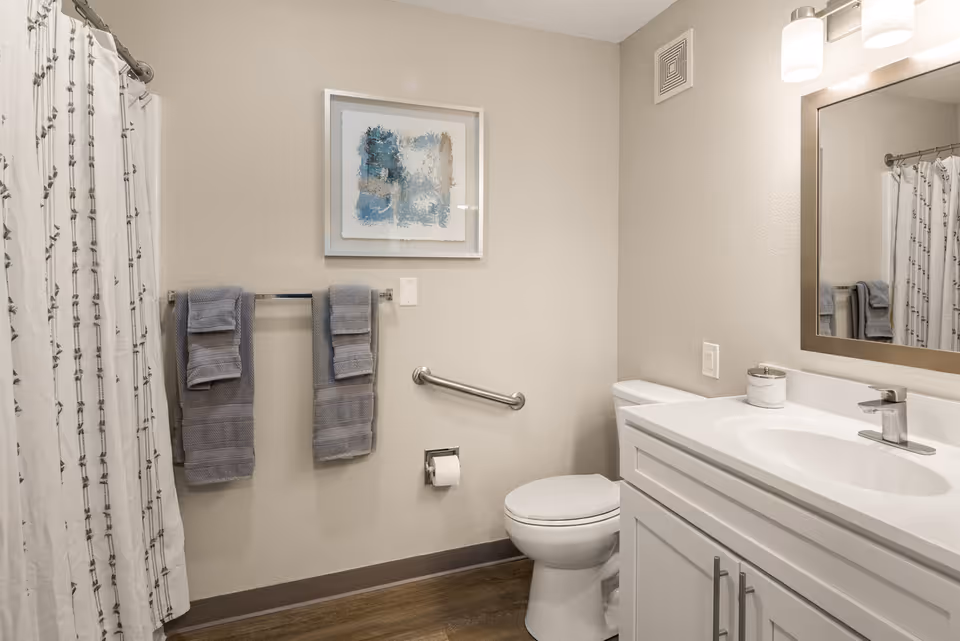 A clean and modern bathroom featuring a white toilet, white vanity with a sink and faucet, a large mirror with two wall-mounted lights above it, a towel rack with two gray towels, a shower curtain with a subtle pattern, and a framed abstract artwork on the wall. The floor has wood-like flooring and the walls are painted light beige.
