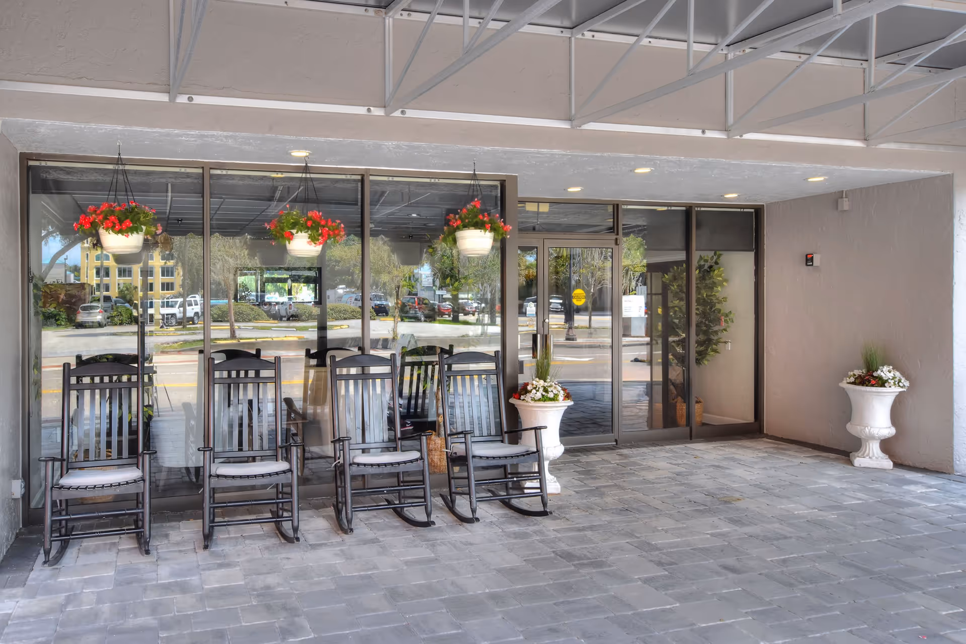 Covered entrance area of a building with four black rocking chairs lined up in front of large glass windows and doors. Three hanging flower pots with red flowers are suspended from the ceiling above the chairs. Two large white planters with flowers are placed on either side of the entrance. The ground is paved with gray bricks.