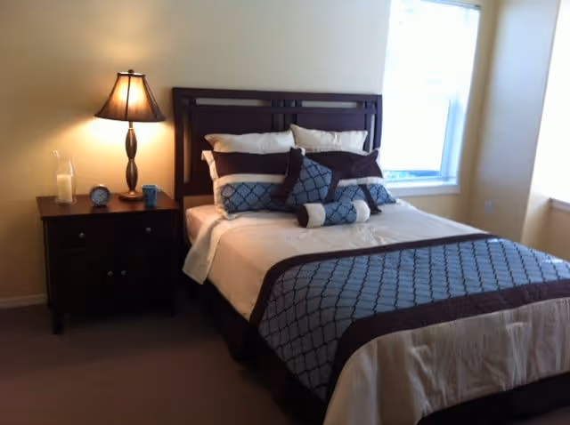 A neatly made bed with multiple pillows and a patterned blue and brown bedspread in a bedroom. Next to the bed is a dark wooden nightstand with a lamp, a clock, and a glass candle holder. There is a window with white blinds on the right side of the room.