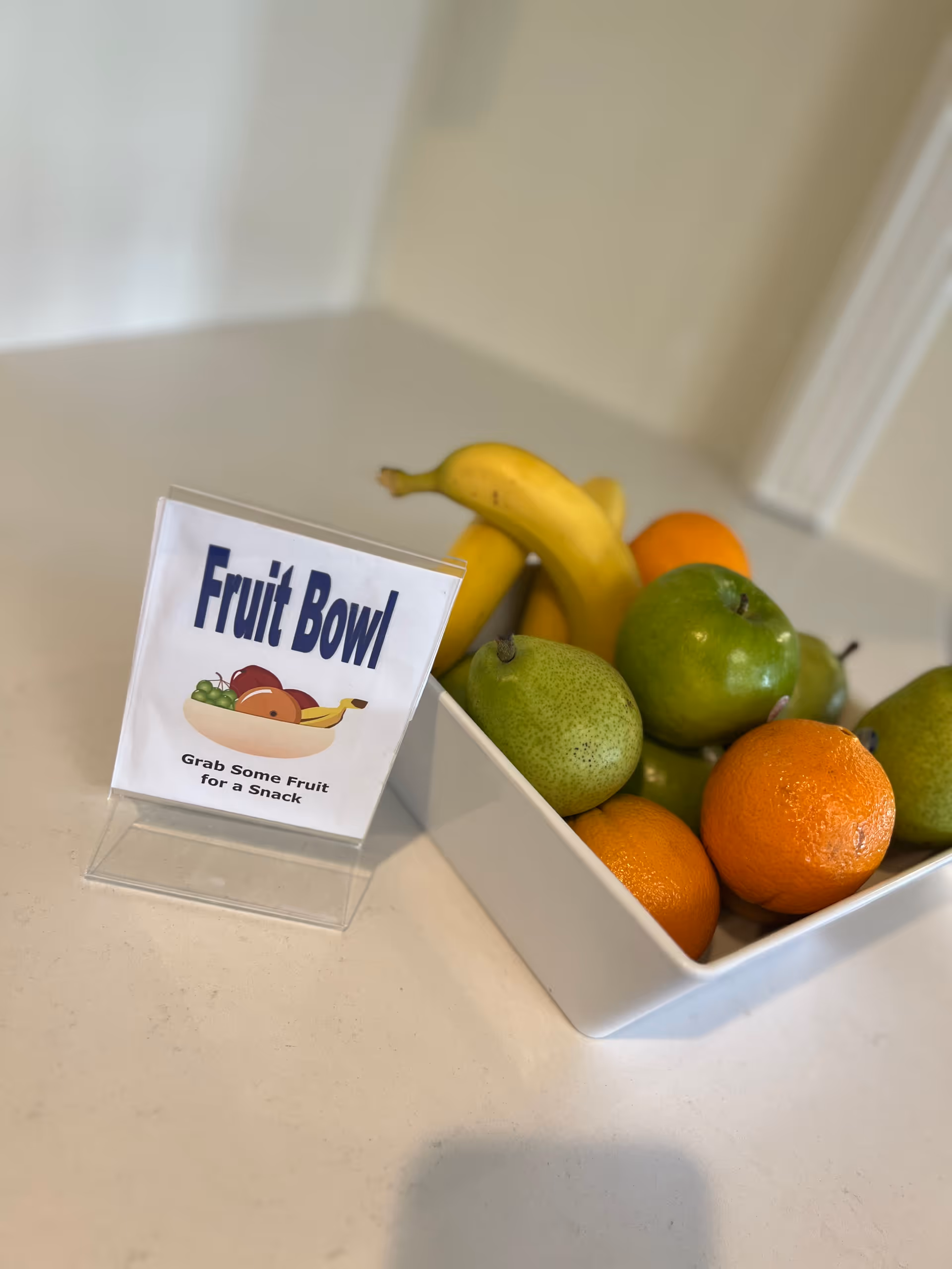White rectangular bowl of assorted fruit (bananas, pears, apples, oranges) on a countertop next to a small sign reading 'Fruit Bowl'.