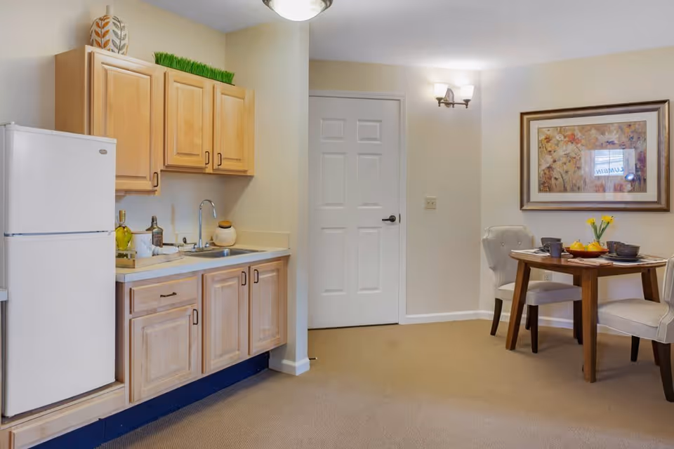 A small kitchen area with light wood cabinets, a white refrigerator, a sink, and countertop with decorative items. Adjacent to the kitchen is a dining area with a round wooden table set for two, two upholstered chairs, a vase with yellow flowers, and a framed floral painting on the wall.