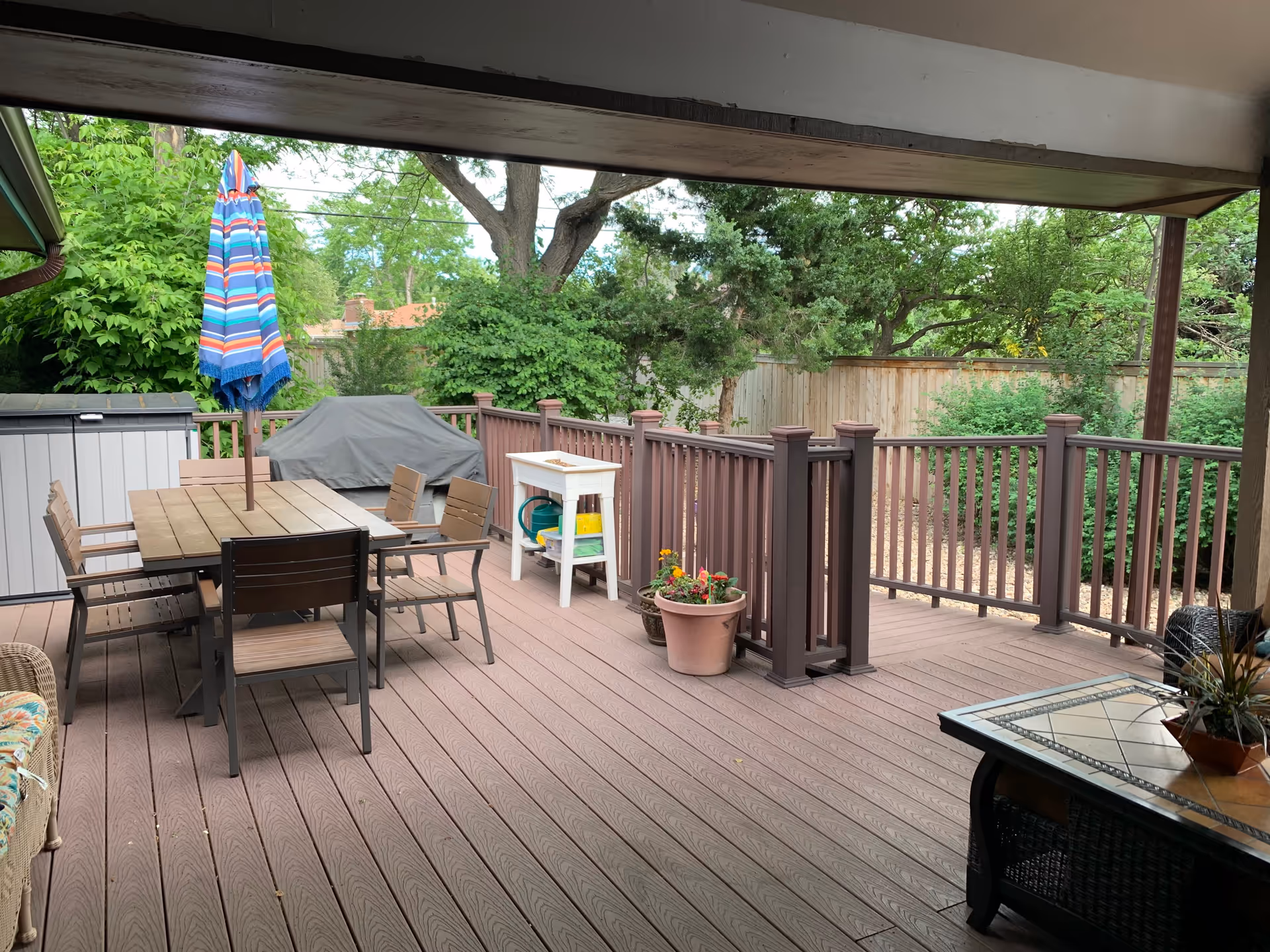 Covered outdoor patio area with a wooden deck, a dining table with six chairs and a closed striped umbrella, a covered grill, potted plants, and a small white table with gardening supplies. The patio is surrounded by a wooden railing and lush green trees and bushes in the background.
