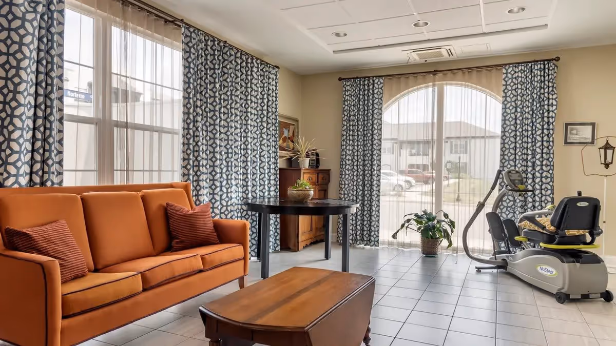 A bright living room with a burnt orange sofa with two matching pillows, a wooden coffee table, a round black table with a plant on it, patterned blue and white curtains covering large windows, a recumbent exercise bike, and a potted plant near the window.