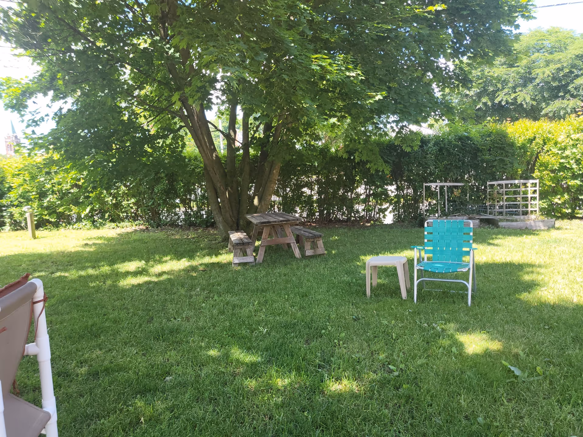 A shaded outdoor garden area with a large tree, a wooden picnic table with benches, a green lawn chair, and a small beige plastic side table on green grass. There are bushes and trees in the background providing privacy and shade.