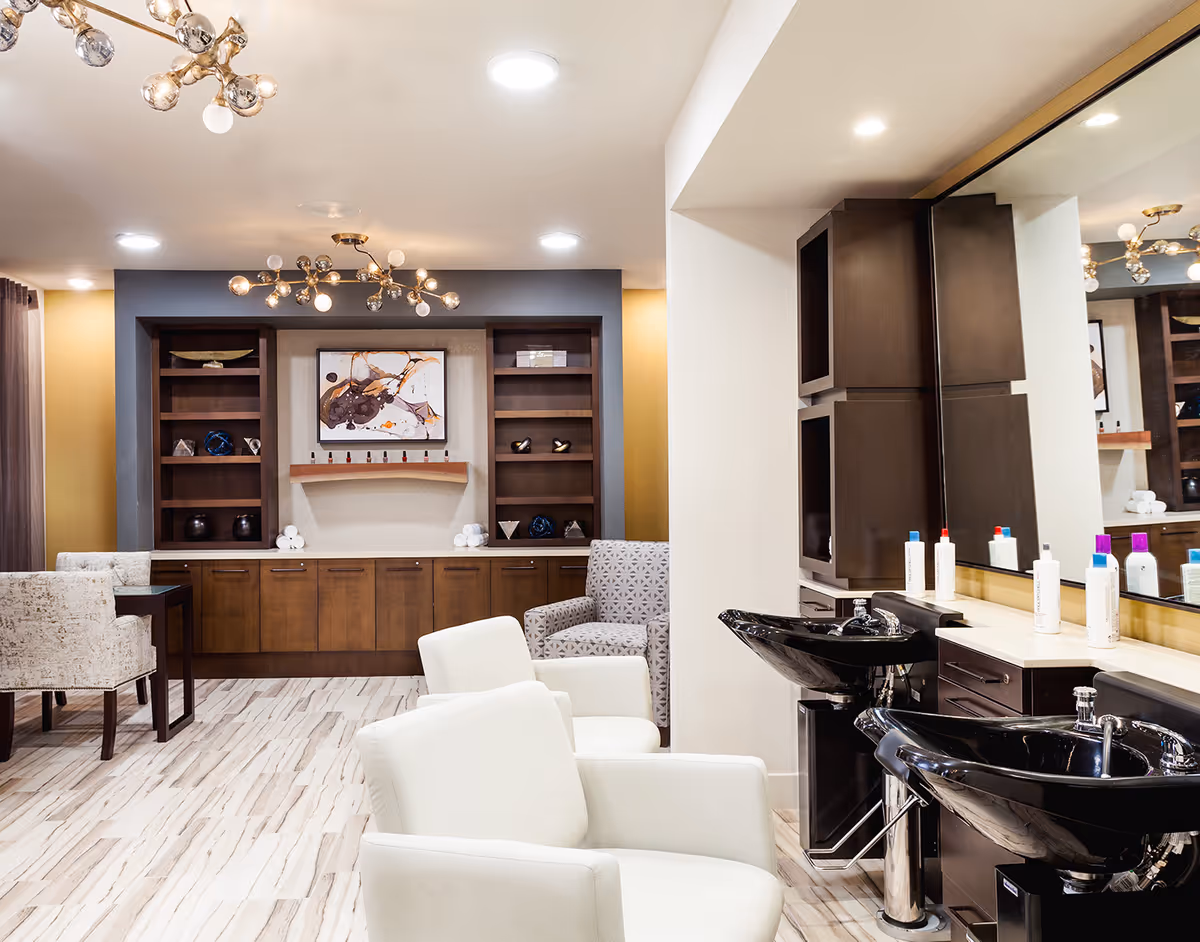 Interior view of a senior living facility's salon area featuring two white salon chairs in front of black wash basins with hair care products on the counter. In the background, there is a seating area with a patterned armchair, a wooden cabinet with shelves, decorative items, and modern light fixtures on the ceiling.
