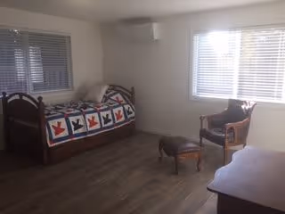 Furnished memory-care bedroom with a single daybed covered by a patchwork quilt, a chair and ottoman, a desk, and two windows.