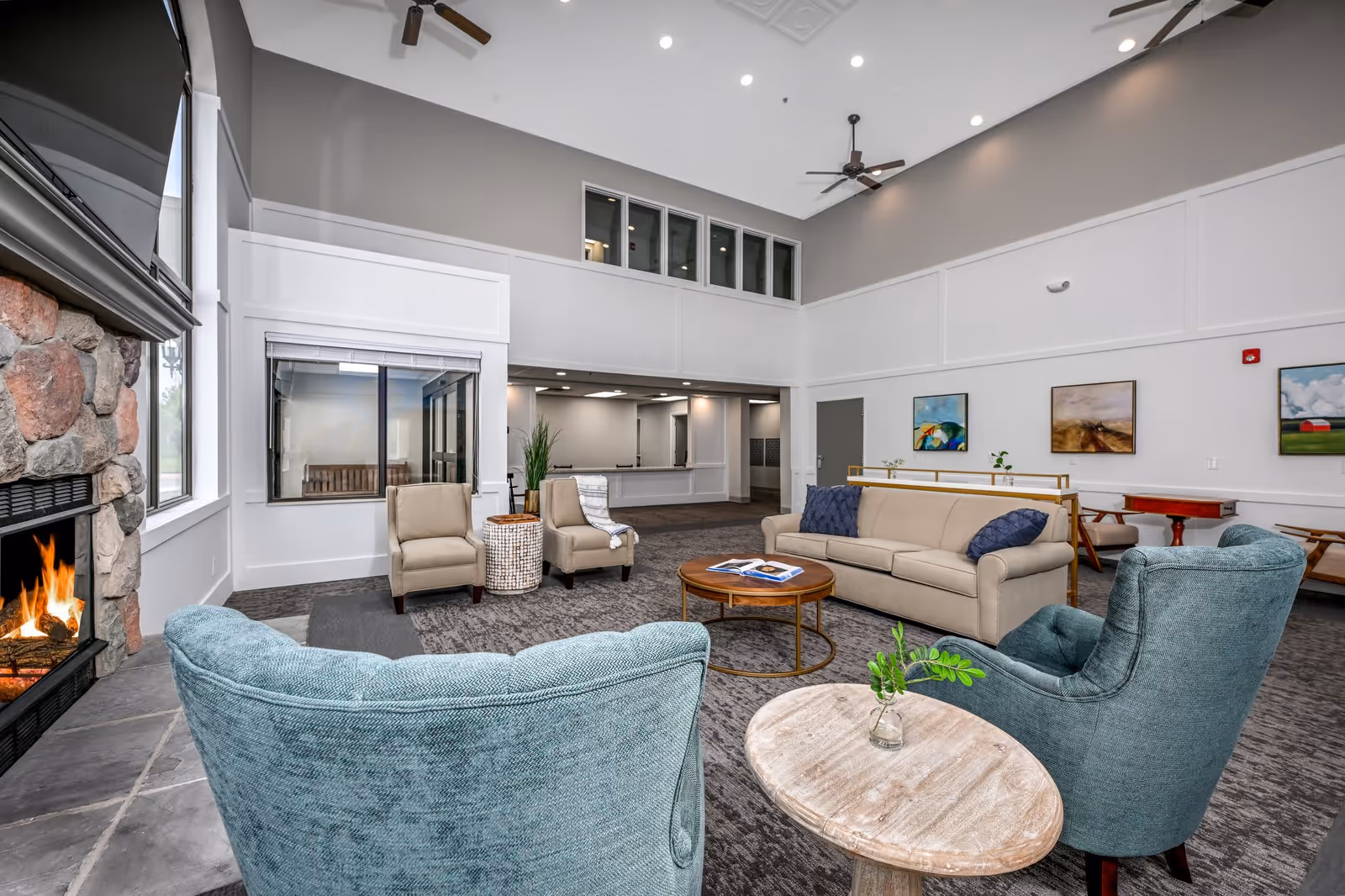 A spacious senior living facility common area with high ceilings, a stone fireplace with a fire burning, two teal armchairs, a beige sofa with blue pillows, two beige armchairs, a round wooden coffee table with an open book, and a small round side table with a small plant. The walls are decorated with three paintings, and there are ceiling fans and recessed lighting.