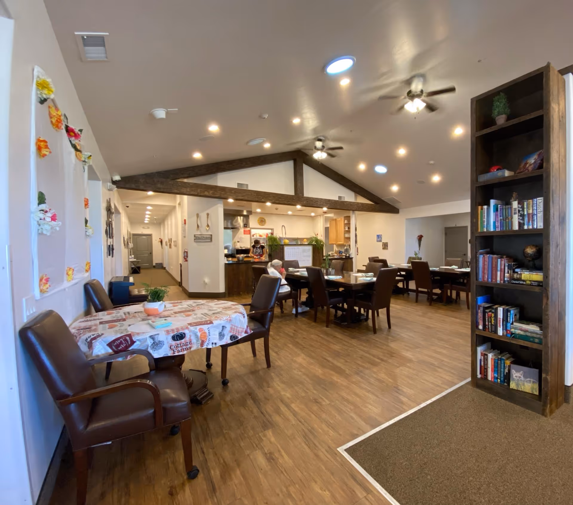 Interior view of a senior living facility dining and common area with wooden floors, several tables and chairs, a bookshelf filled with books, ceiling fans, and recessed lighting. A person is seated at one of the tables, and a staff member is visible near the kitchen area in the background.