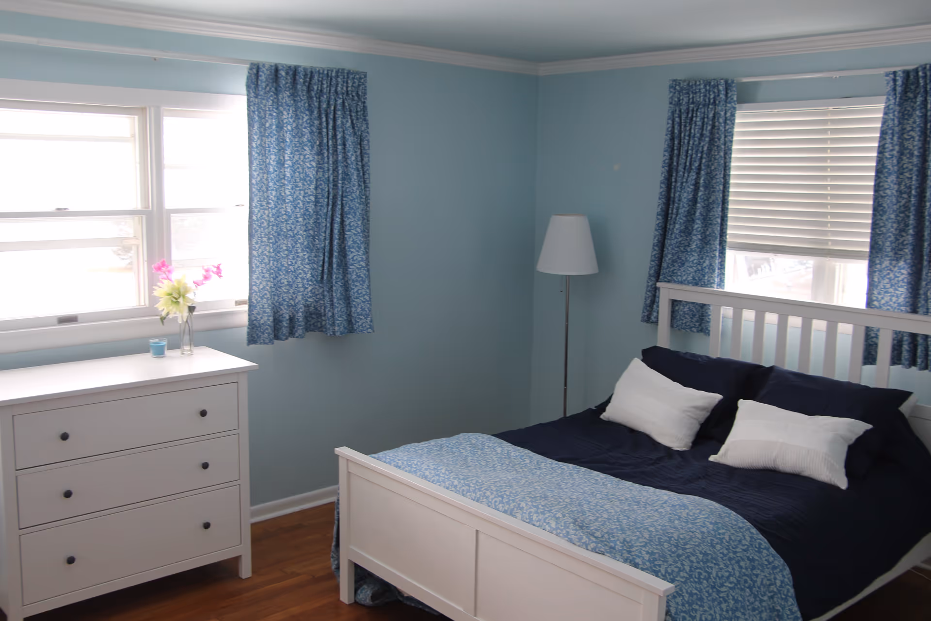 A bedroom with light blue walls and two windows covered with blue patterned curtains. There is a white wooden bed with dark blue bedding and two white pillows. A white dresser with three drawers is placed under one window, decorated with a small vase of flowers and a candle. A tall white floor lamp stands in the corner near the bed.