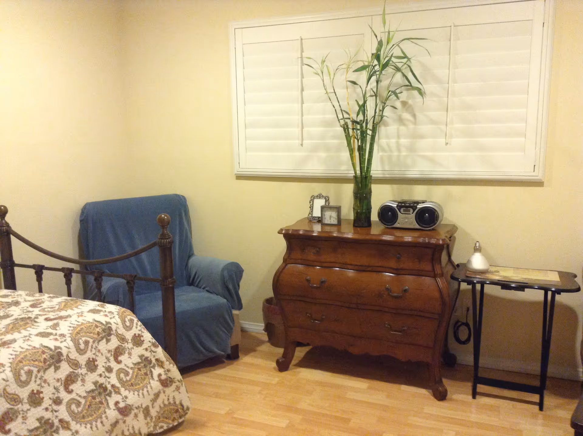 A cozy bedroom corner with a wooden bed frame partially visible, a blue armchair covered with a slipcover, a wooden dresser with three drawers, a vase with tall green plants, a small clock, a picture frame, and a radio on top. There is also a small side table with a lamp and a decorative item. The room has light-colored walls and a window with white shutters.