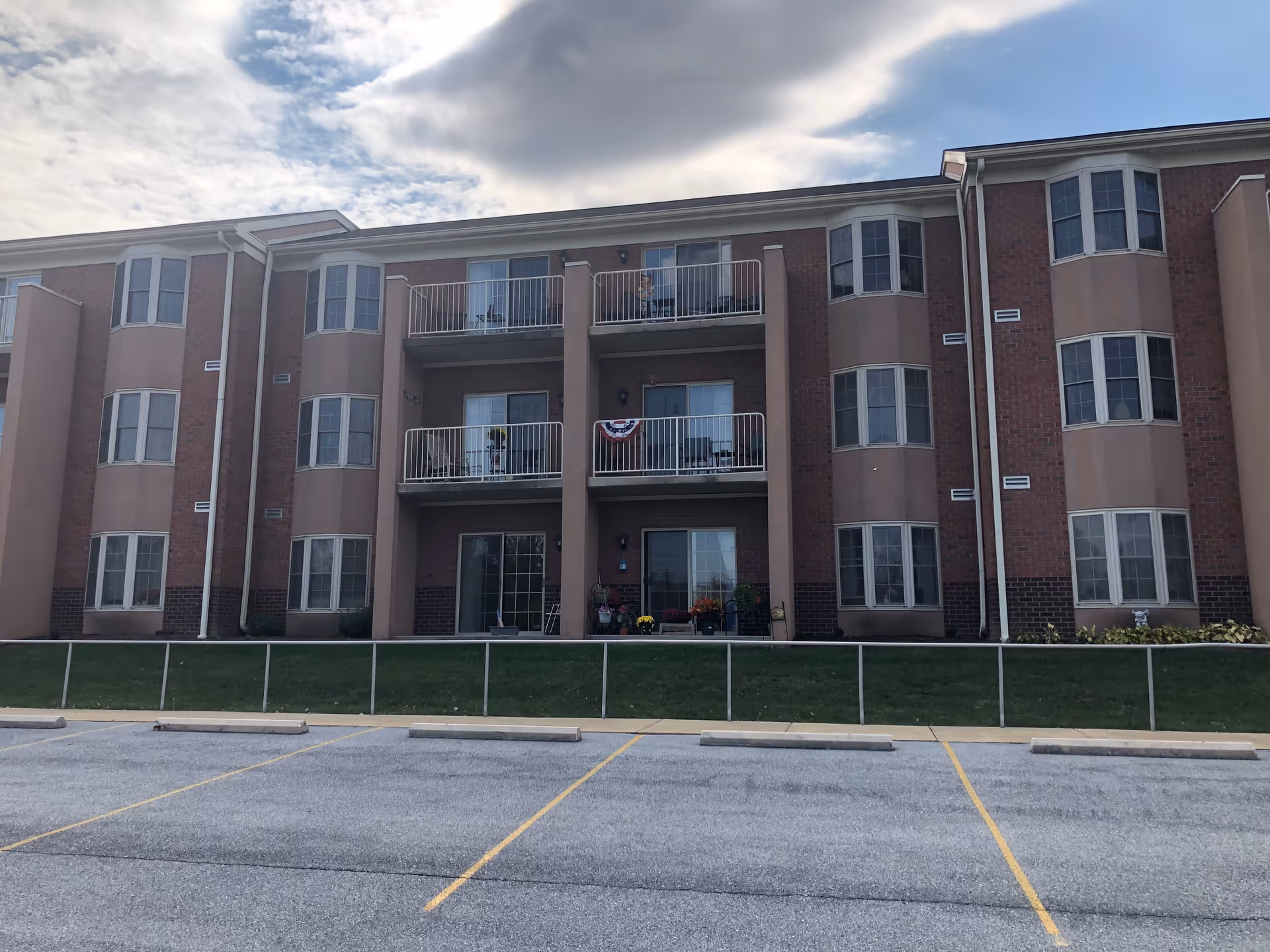Exterior view of a three-story senior living facility building with balconies on the second and third floors. The building has a brick and beige facade with multiple windows. There is a parking lot with marked spaces in the foreground and a grassy area with a metal railing between the building and the parking lot. The sky is partly cloudy.