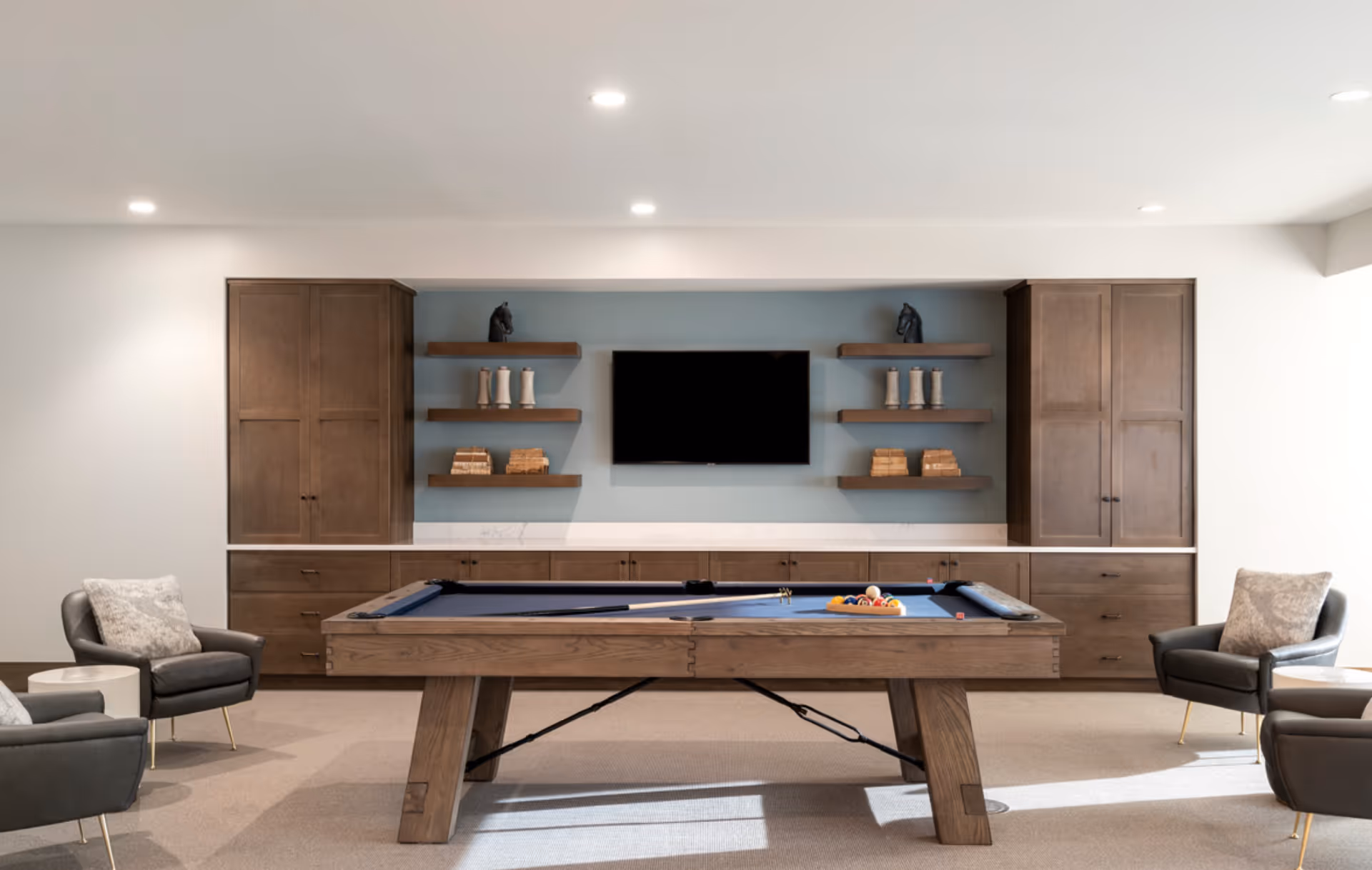 A modern game room with a pool table in the center, flanked by four gray armchairs with cushions. The back wall features built-in wooden cabinets and shelves with decorative items, and a flat-screen TV mounted in the middle. The room is well-lit with recessed ceiling lights.
