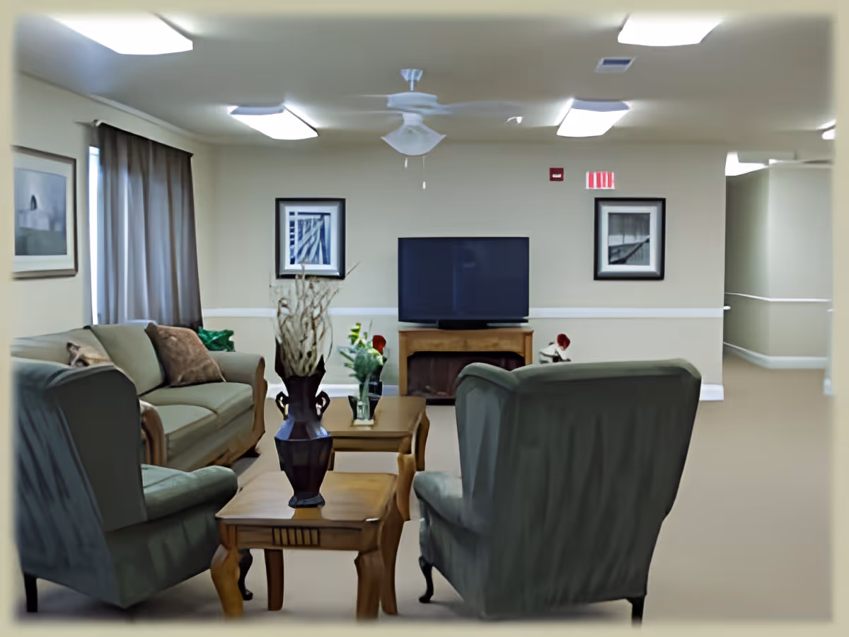 A cozy living room with two armchairs and a sofa arranged around two wooden coffee tables. The tables have decorative vases with flowers and branches. A flat-screen TV is mounted on a wooden stand against the far wall, which is decorated with two framed pictures. The room has beige walls, a ceiling fan, and overhead fluorescent lighting. A window with brown curtains is on the left side.