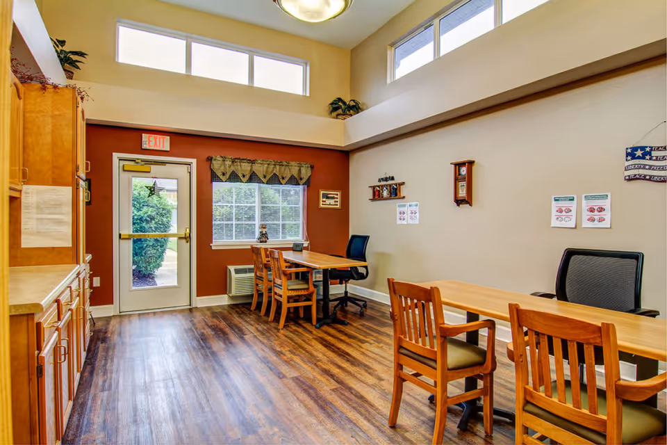 Interior room with wooden flooring, two tables each with chairs and office chairs, a window with a valance, an exit door, and cabinets on the left side. The walls are painted beige and brown, with some wall decorations and informational posters.
