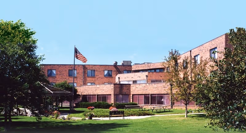 Exterior view of a multi-story brick building with several windows, surrounded by a well-maintained lawn, trees, and bushes. There is a flagpole with an American flag in front of the building, along with benches and picnic tables on the grass under a clear blue sky.
