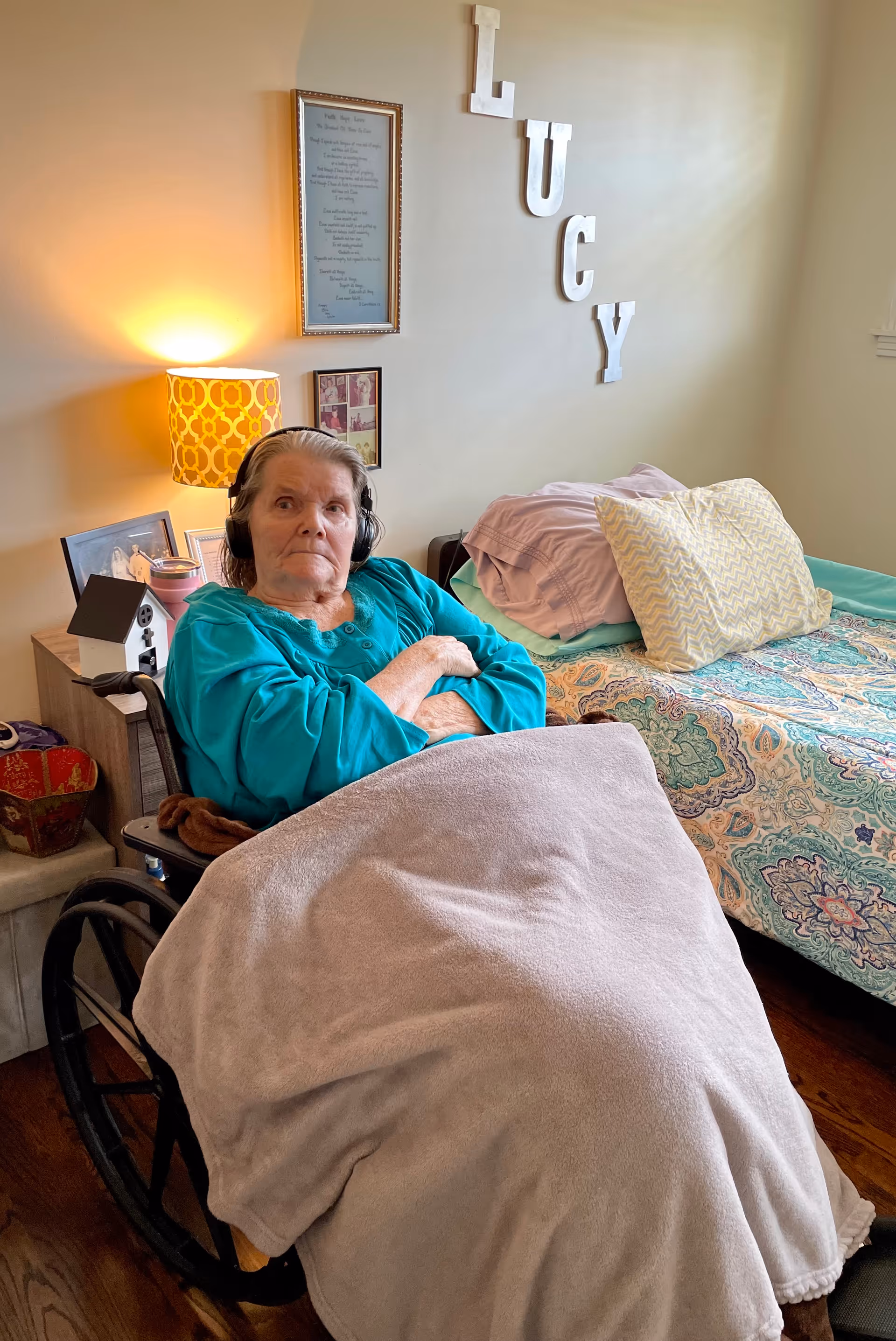 An elderly woman sitting in a wheelchair covered with a beige blanket in a bedroom. She is wearing a teal top and headphones. Behind her is a bed with colorful bedding and pillows, a nightstand with a lamp, framed pictures on the wall, and large white letters spelling 'LUCY'.