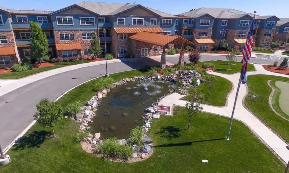 Exterior view of Keystone Place At Legacy Ridge, showing a three-story building with a stone and blue facade, a covered entrance, a landscaped area with a pond and fountain, benches, trees, and an American flag on a flagpole.
