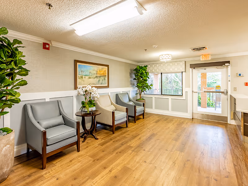 A bright and clean waiting area with three armchairs arranged along a wall, a small round table with a flower arrangement, two large potted plants, a framed landscape painting, and a glass door leading outside. The floor is wooden, and the walls are light-colored with white trim.