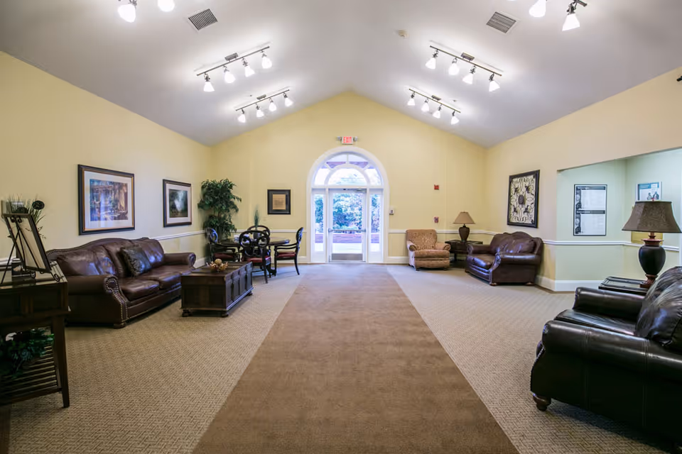 A spacious, well-lit common area with beige walls and carpeted floors. The room features several leather sofas and armchairs arranged around wooden coffee tables. There are framed pictures on the walls, a potted plant, and a small round table with chairs near the back. Large glass double doors with an arched window above lead outside, allowing natural light to enter the room.