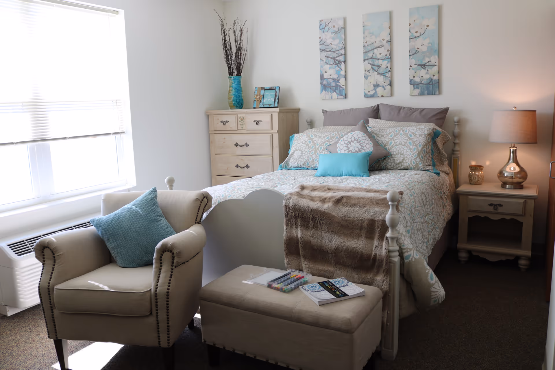 A cozy bedroom with a bed adorned with patterned bedding and multiple pillows in shades of blue and gray. A beige armchair with a blue cushion sits near a window with blinds. At the foot of the bed is a beige ottoman with coloring markers and a book on top. A wooden dresser with a blue vase and picture frame is against the wall, and a nightstand with a lamp and candle is beside the bed. Three floral paintings hang above the bed on a white wall.