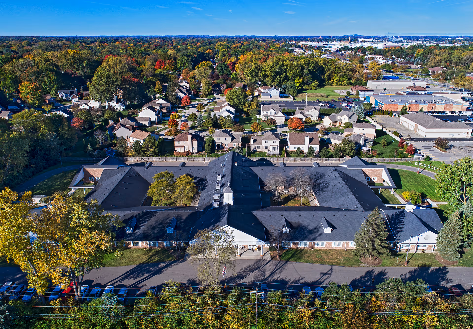 Aerial view of American House Livonia facility surrounded by trees and residential houses, with a parking area in front and a clear blue sky above.
