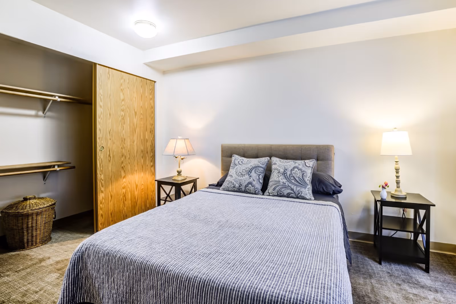 A bedroom with a neatly made bed featuring a gray upholstered headboard and two patterned pillows. There are two black nightstands on either side of the bed, each with a lamp. To the left, there is an open closet with wooden sliding doors and a wicker basket inside. The room has beige carpeting and white walls.