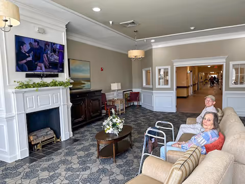 A cozy living room area in an assisted living facility with a beige couch where an elderly man and woman are seated. The room features a fireplace with a TV mounted above it, a wooden coffee table with a flower arrangement, patterned carpet, and a hallway visible in the background.