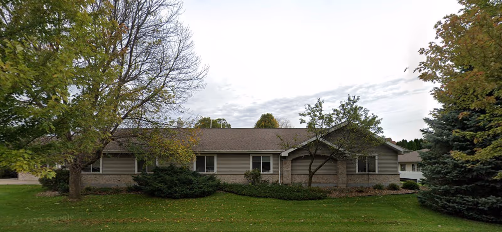 Single-story building with beige siding and a brown roof surrounded by green grass, trees, and shrubs under a cloudy sky.