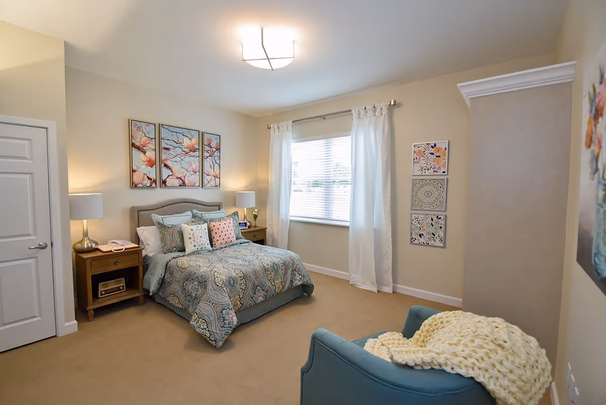A bright, softly decorated bedroom with a patterned bed, bedside tables and lamps, a window with curtains, and an armchair draped with a blanket.