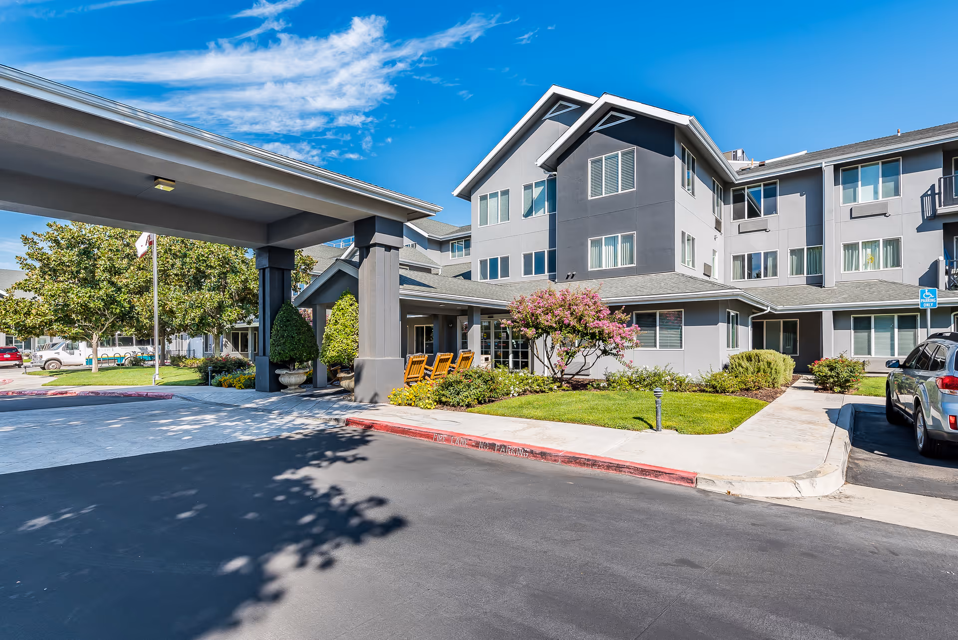 Exterior view of Solstice Senior Living at Clovis, showing a modern three-story building with gray walls and multiple windows. The entrance features a covered drop-off area with columns, surrounded by well-maintained landscaping including green grass, bushes, and a flowering tree. There are parked cars and a clear blue sky above.