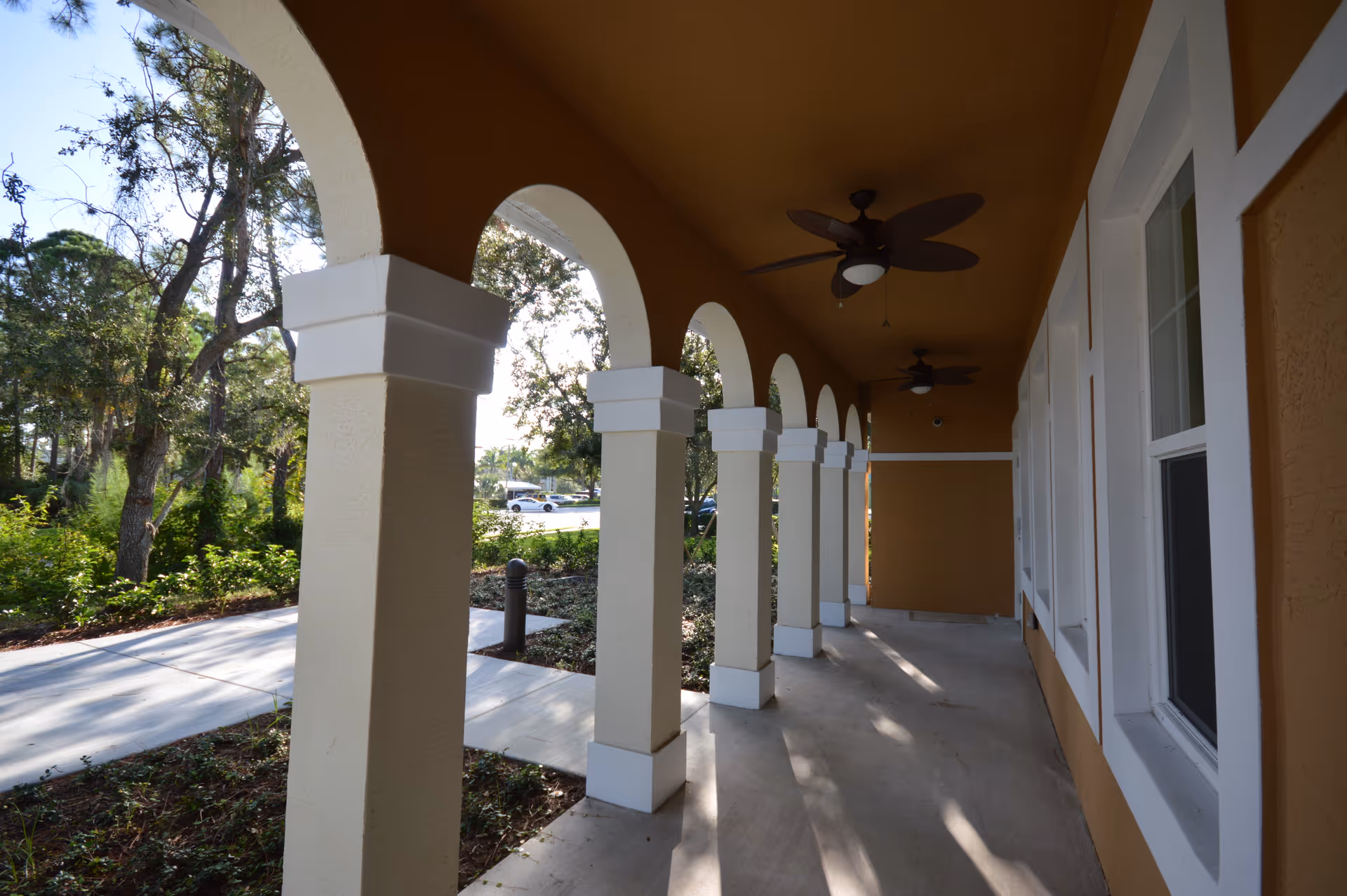 Covered outdoor walkway with beige columns and arches, ceiling fans, and windows on the right side. Trees and greenery are visible outside along with a sidewalk and parked cars in the distance.