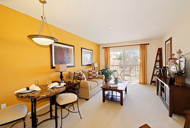 Bright and cozy living room with a yellow accent wall, a small round dining table set for two with wine glasses and napkins, a beige sofa with patterned cushions, a wooden coffee table, a TV stand with decorative items, and a sliding glass door leading to a balcony with outdoor seating.