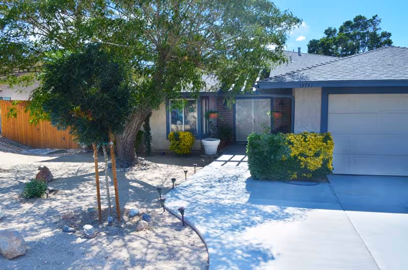 Front exterior of a single-story residence with a driveway and garage, shaded walkway, trees and shrubs.