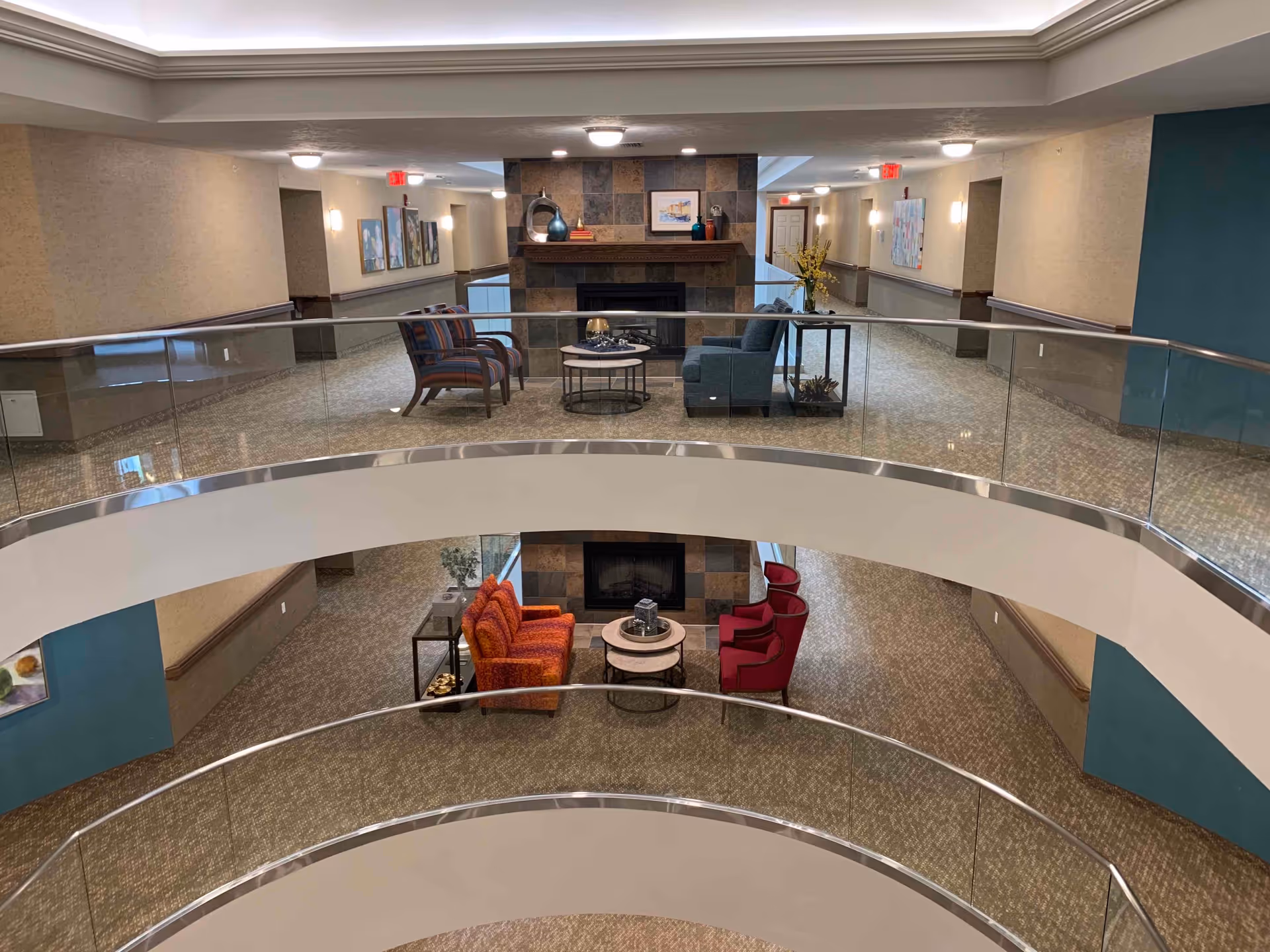 Interior view of a multi-level common area in a senior living facility featuring seating arrangements with chairs and tables around a fireplace on both levels, with glass railings and carpeted floors.