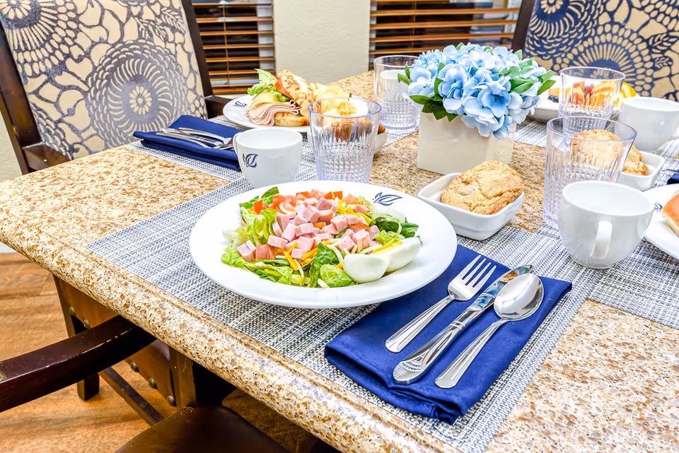 A dining table set with plates of salad topped with diced ham and hard-boiled egg halves, glasses of water, cups, muffins in small dishes, and a centerpiece of blue flowers. The table has a granite surface with placemats and blue cloth napkins with silverware. Upholstered chairs with floral patterns are visible around the table.