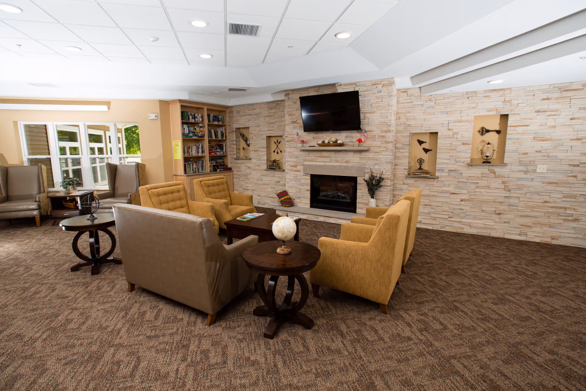 A cozy living room area with a stone fireplace and a mounted flat-screen TV above it. The room features a mix of beige and mustard yellow armchairs arranged around a dark wooden coffee table and side tables. There is a bookshelf filled with books in the corner and decorative items displayed in recessed wall niches. Large windows on the left side let in natural light, and the carpet has a patterned design.
