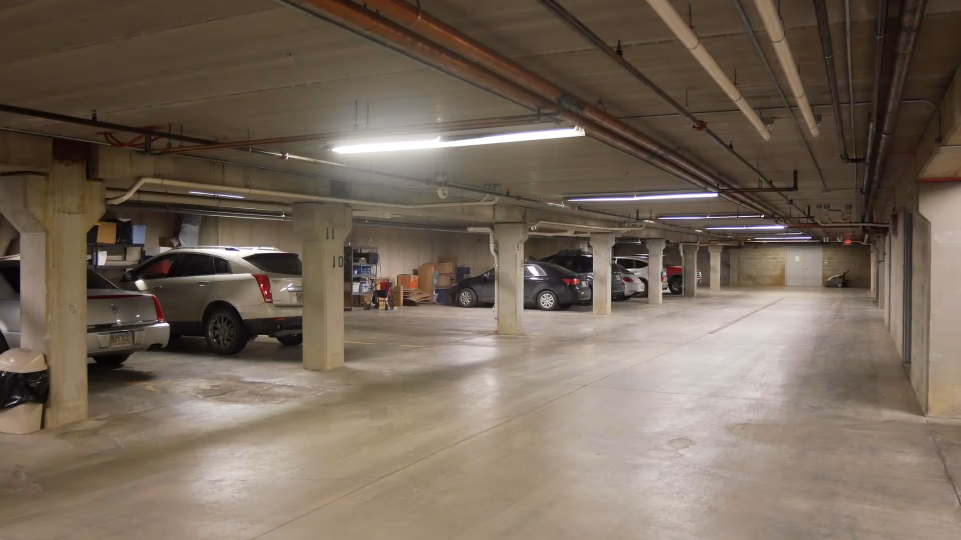 Underground parking garage with concrete pillars, several parked cars, and fluorescent ceiling lights.