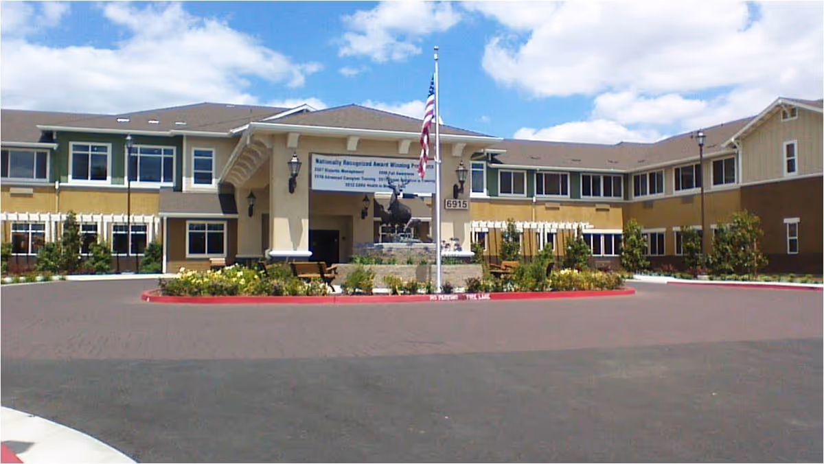Front exterior view of Carlton Senior Living Elk Grove building with a circular driveway, landscaped garden area with benches, an American flag on a flagpole, and a statue in the center under a partly cloudy sky.
