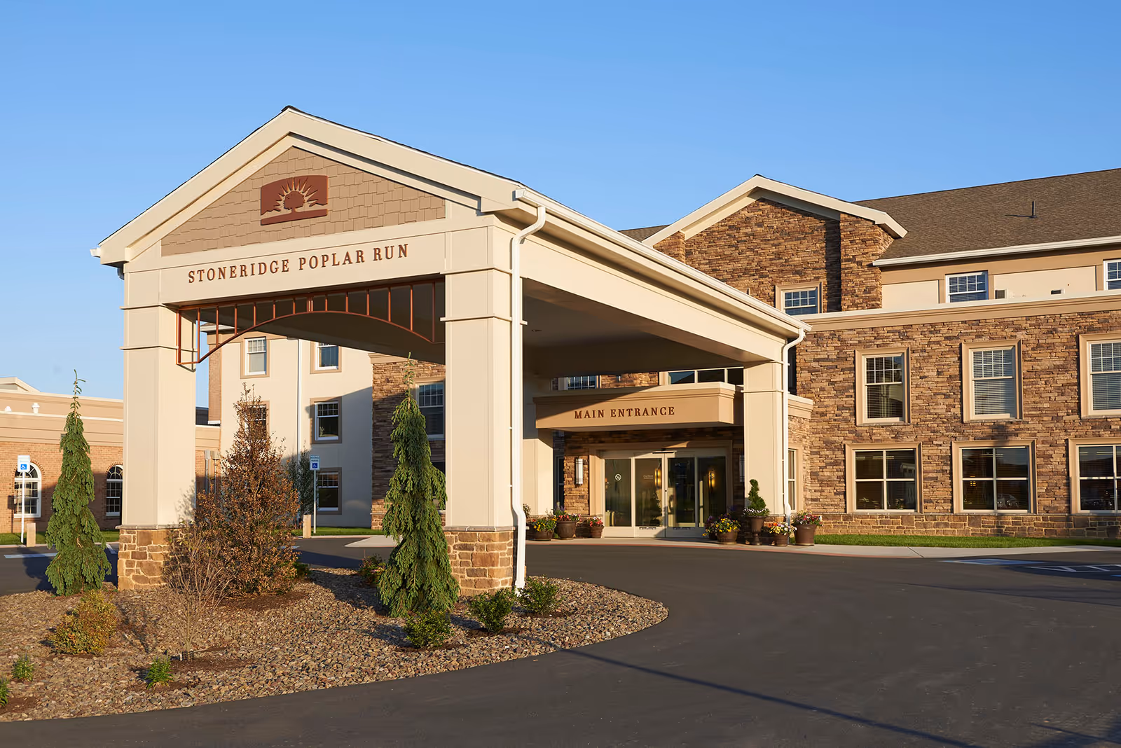 Exterior view of the main entrance of Stoneridge Poplar Run, a retirement living community. The building features a covered driveway with pillars and a sign displaying the community name. The facade is made of stone and beige siding, with several windows and landscaped greenery around the entrance.