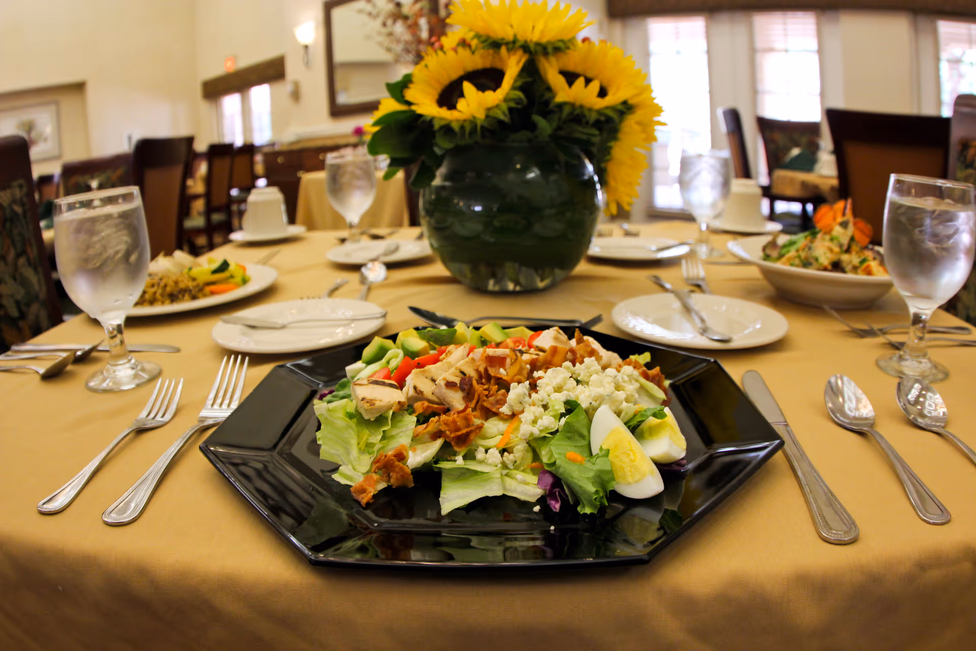 A dining table set with plates of salad, glasses of water, and silverware in a well-lit dining room. A vase with bright yellow sunflowers is placed in the center of the table. The background shows more tables and chairs in the dining area.