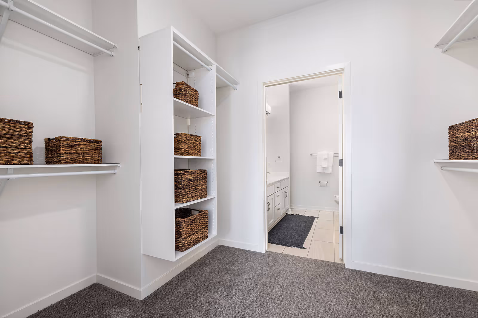 A spacious walk-in closet with white walls and carpeted floor, featuring multiple shelves and hanging rods with woven baskets placed on the shelves. The closet opens into a bathroom with white cabinetry, a dark rug on the tiled floor, and white towels hanging on a towel rack.
